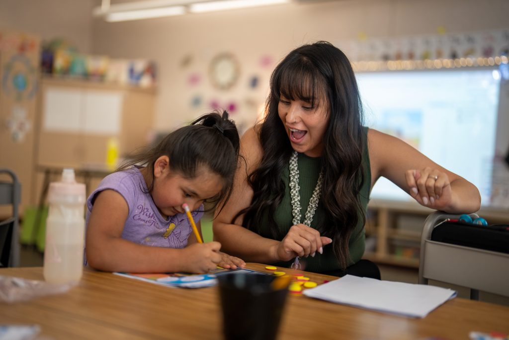Una profesora y una niña realizan una actividad creativa en una mesa; ambas sonríen y están concentradas en su trabajo.