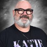 A smiling man with glasses and a beard, wearing a black shirt with "KATIE" printed on it, against a gray background.