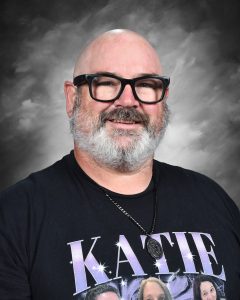 A smiling man with glasses and a beard, wearing a black shirt with "KATIE" printed on it, against a gray background.