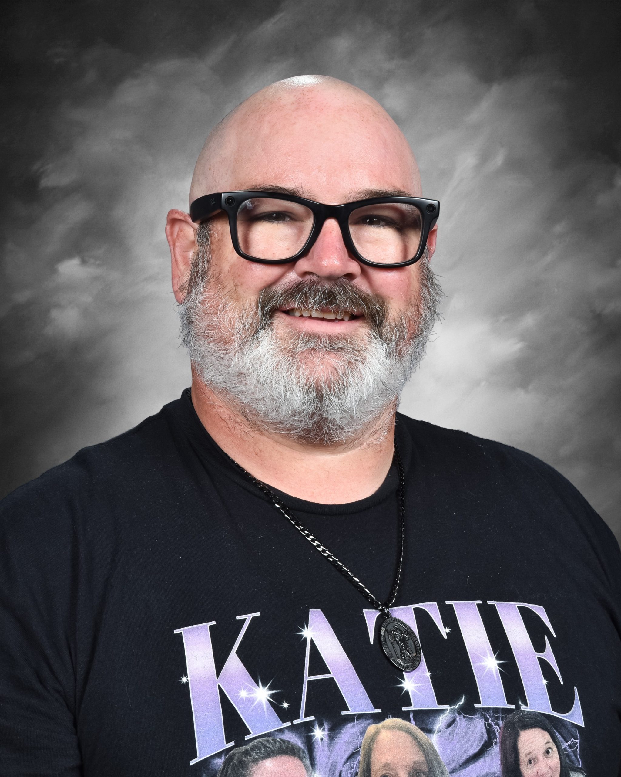 A smiling man with glasses and a beard, wearing a black shirt with "KATIE" printed on it, against a gray background.