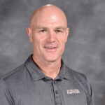 A bald man smiles, wearing a gray polo shirt with "Silver Creek Raptors" logo, against a gray background.