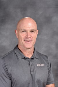A bald man smiles, wearing a gray polo shirt with "Silver Creek Raptors" logo, against a gray background.