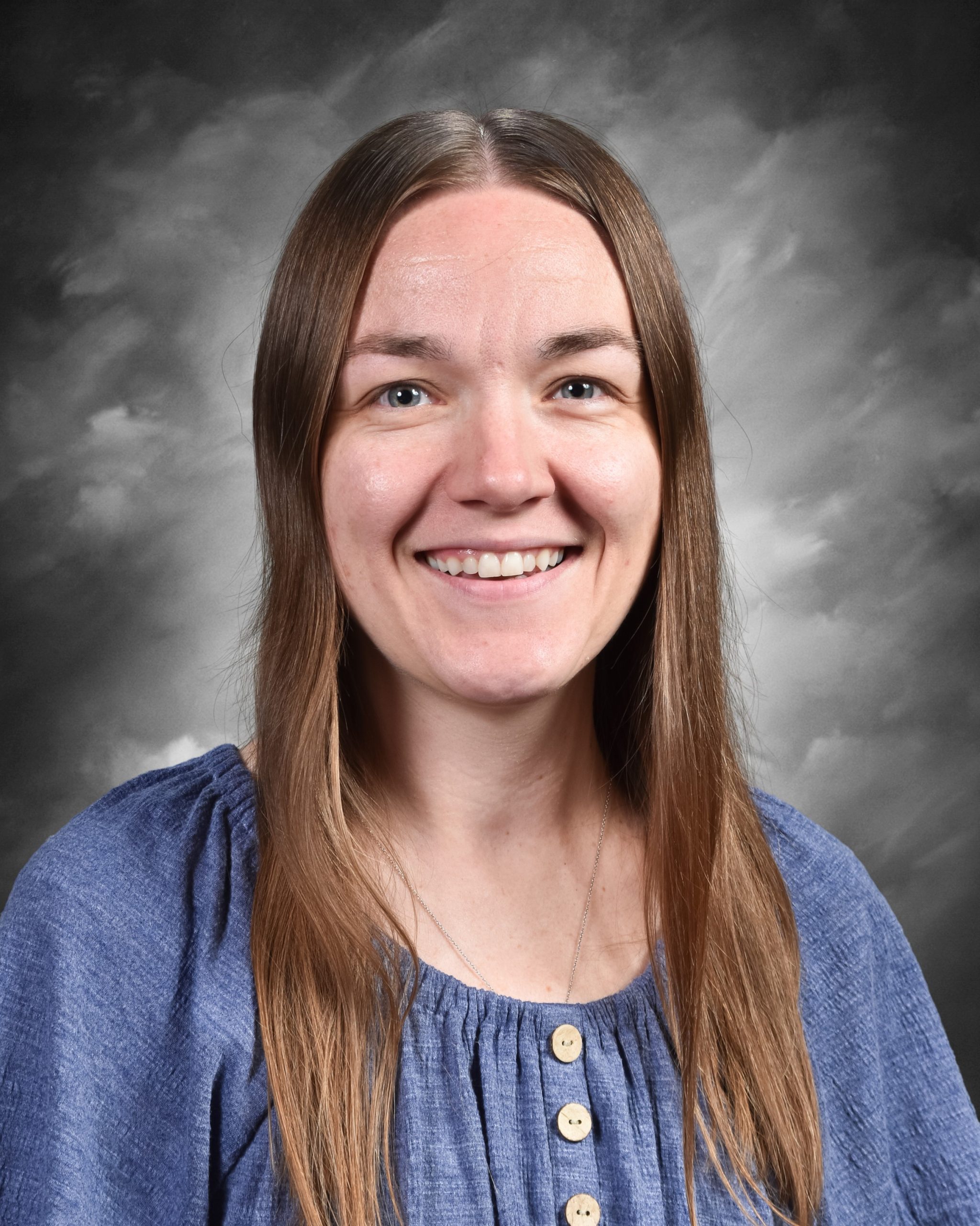 A person with long hair smiles at the camera, wearing a blue blouse with buttons, against a cloudy gray background.