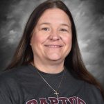 A woman with long brown hair wearing a "Raptor" shirt and a necklace, smiling against a gray background.