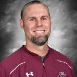 Smiling man in a maroon baseball jacket with a gray background.