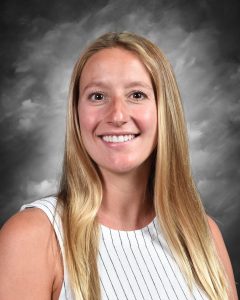 Smiling woman with long blonde hair, wearing a sleeveless striped top against a gray, cloudy background.