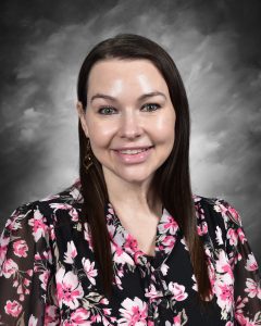 A woman with long brown hair wearing a floral blouse, smiling, against a gray background.