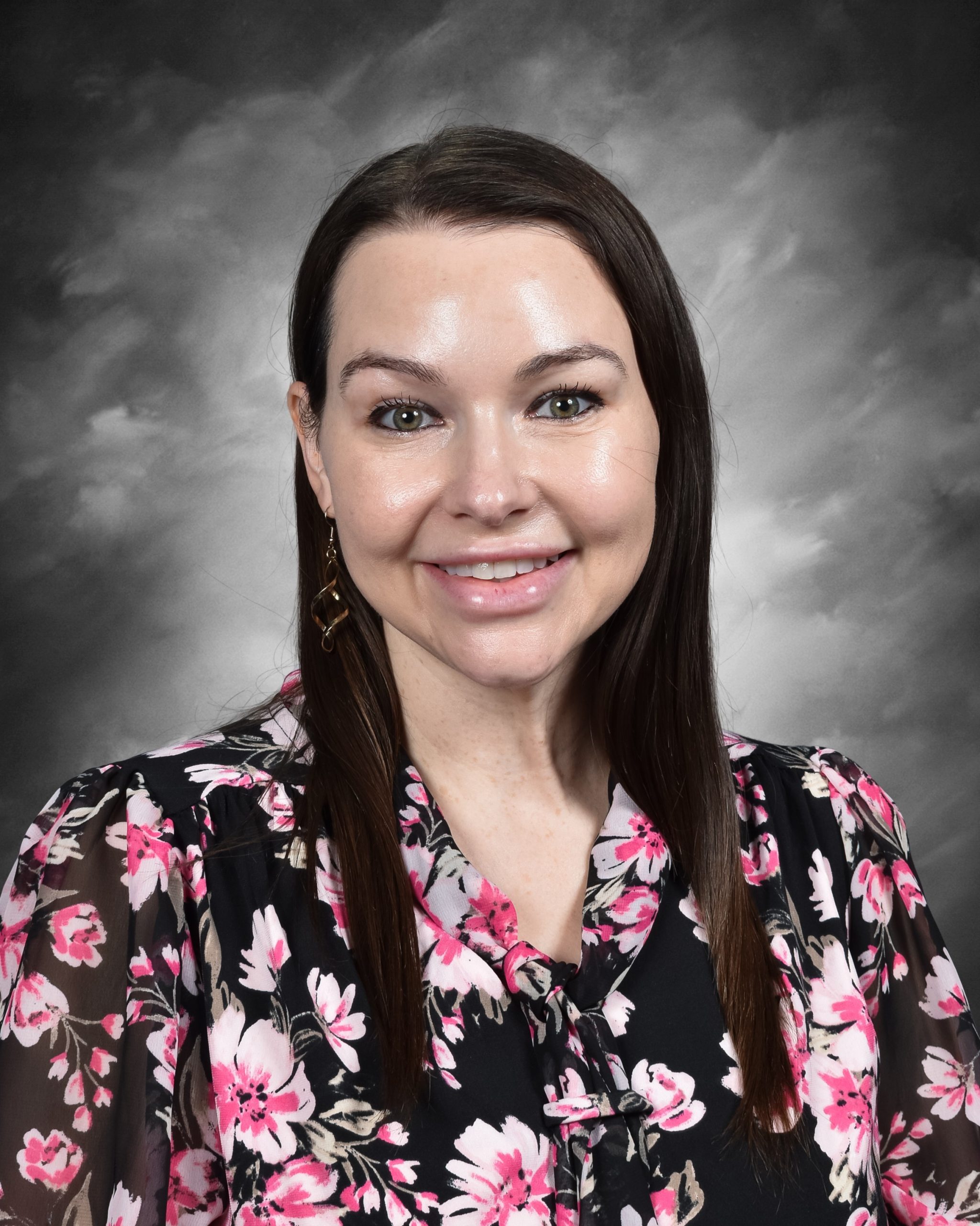 A woman with long brown hair wearing a floral blouse, smiling, against a gray background.