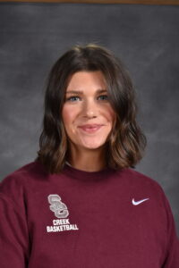 A woman with shoulder-length brown hair smiles, wearing a maroon sweatshirt with "Creek Basketball" logo.
