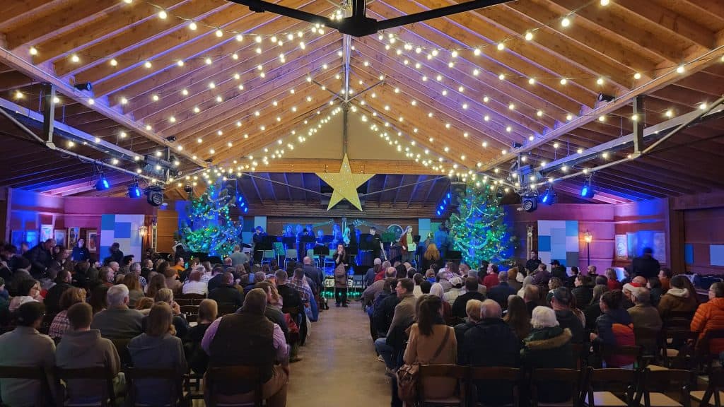 View from back of room looking at stage at Planet Bluegrass. Audience members are seated in chairs and a band is on stage about to perform. Strands of Edison lights are hanging in swags and rows from the ceiling. A giant star is hanging from ceiling in the center.  