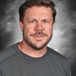 Man with short hair and a beard wearing a dark t-shirt, set against a cloudy gray background.