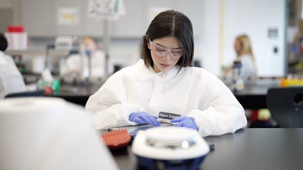 A student in protective gear works focused at a lab table, with equipment and classmates visible in the background.