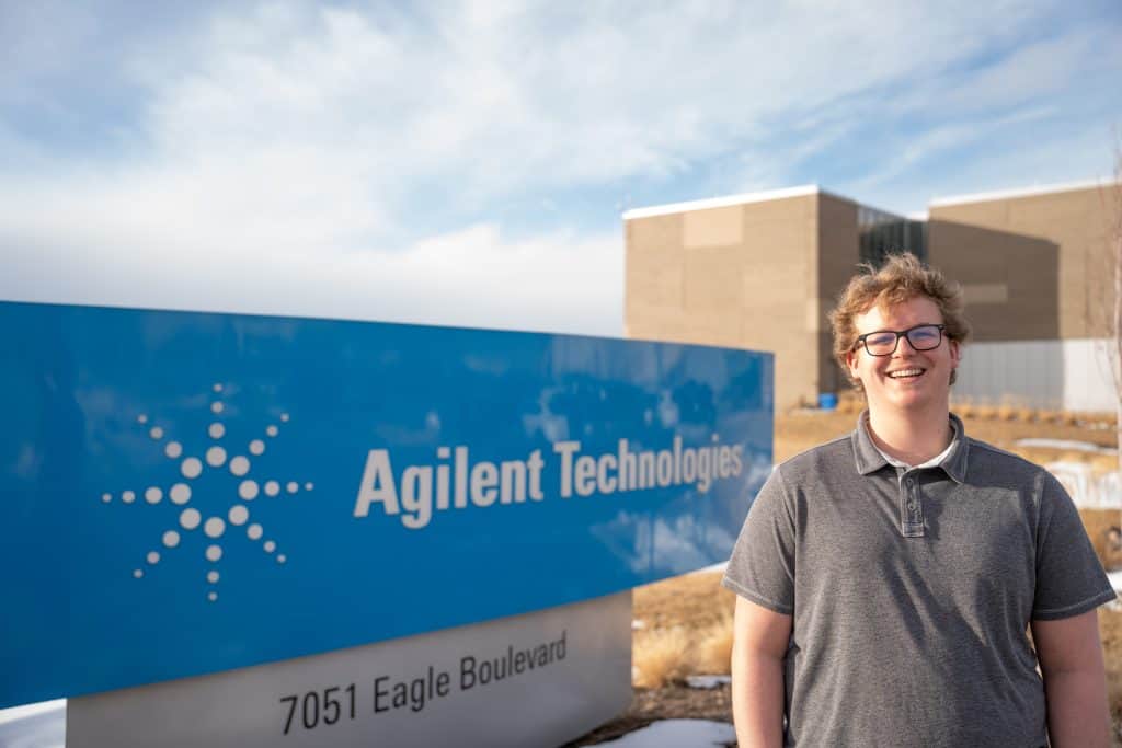 A smiling young man stands in front of the Agilent Technologies sign on a sunny day.