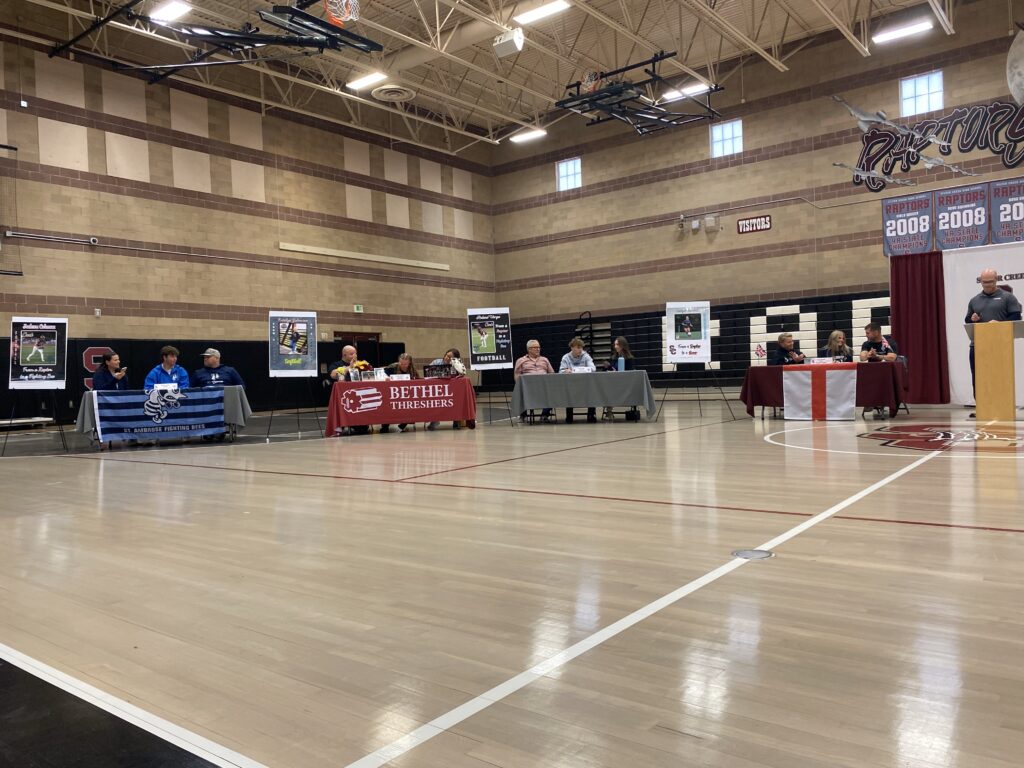 A gymnasium event featuring multiple tables with participants, banners, and a speaker at a podium.