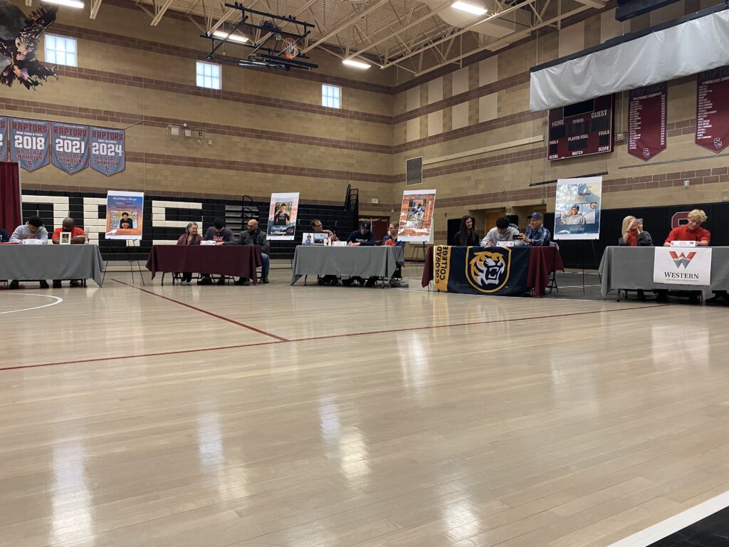 A gymnasium with multiple tables set up for an event, featuring college banners and individuals seated at the tables.