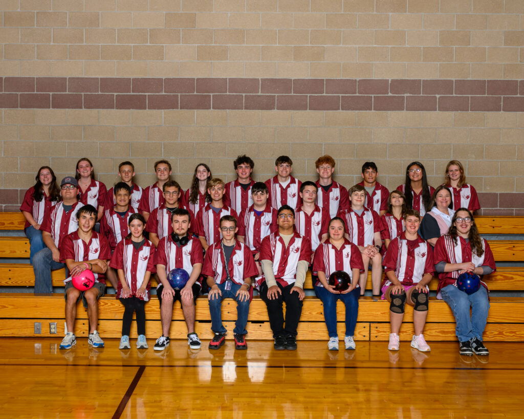 A group of 33 students wearing red and white bowling shirts poses on bleachers in a gymnasium.