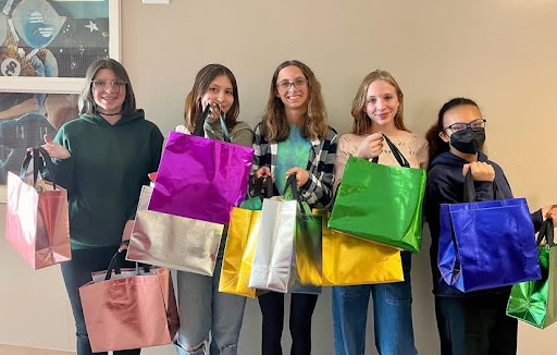 Five students stand together, each holding colorful shopping bags, smiling in a brightly lit indoor space.