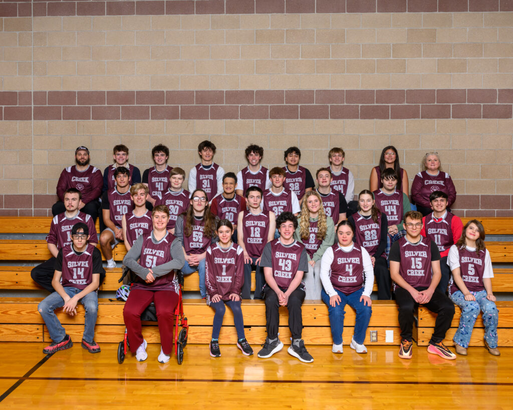 Group photo of a diverse basketball team wearing maroon jerseys, seated on wooden bleachers in a gymnasium.